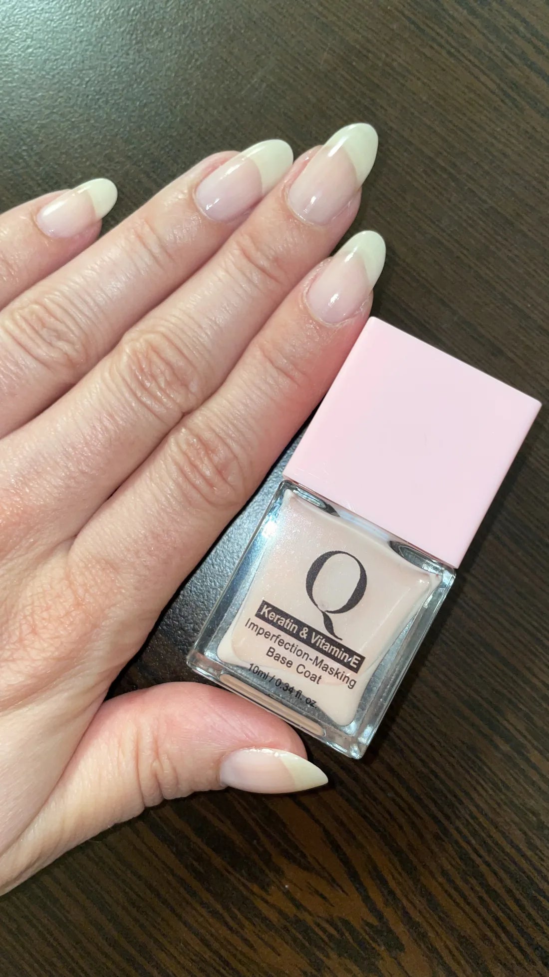 A hand with natural nails wearing one coat of the sheer pink Imperfection-Masking Base Coat, the bottle resting in the palm